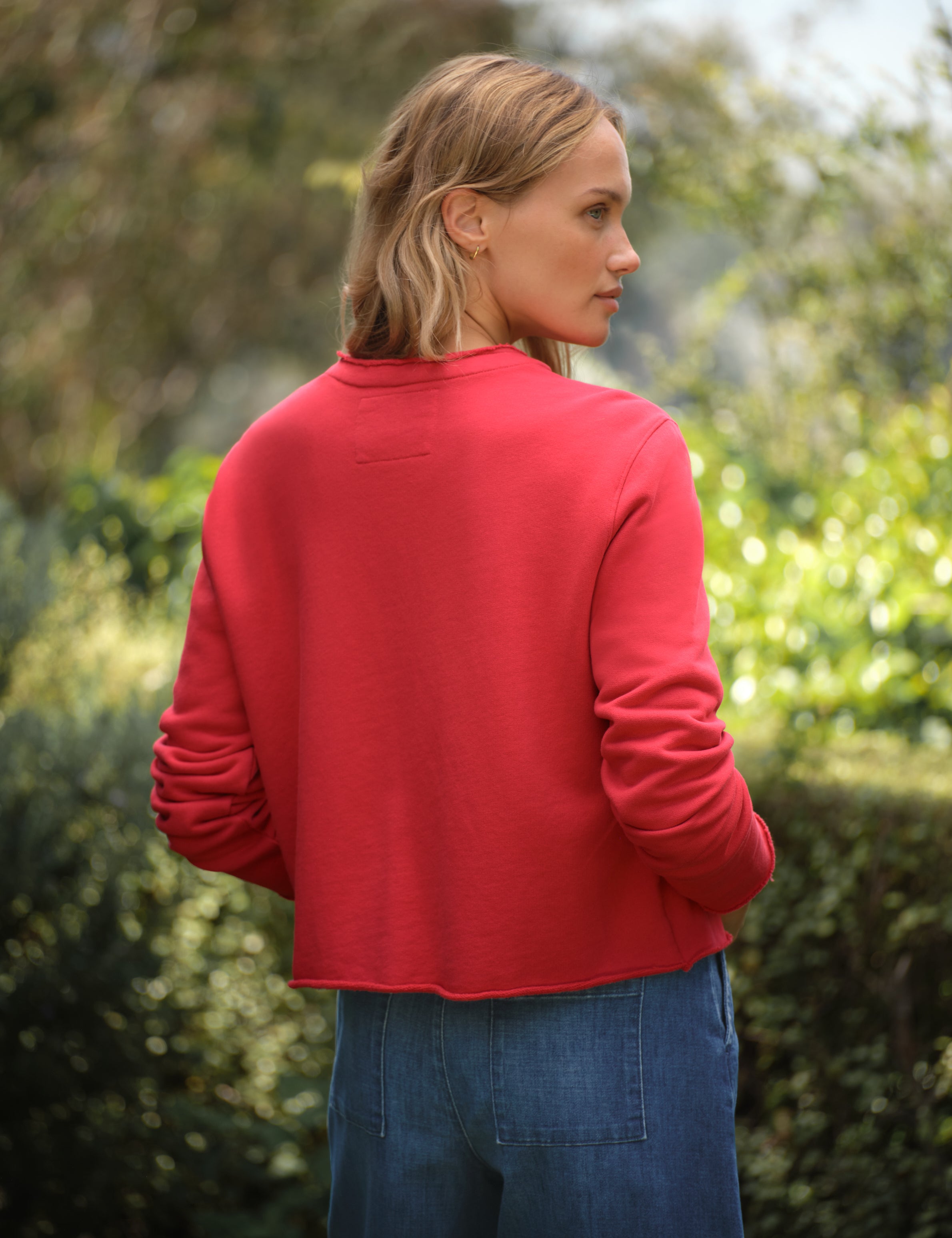 Person wearing a red cardigan and blue jeans standing outdoors with greenery in the background, alternate view