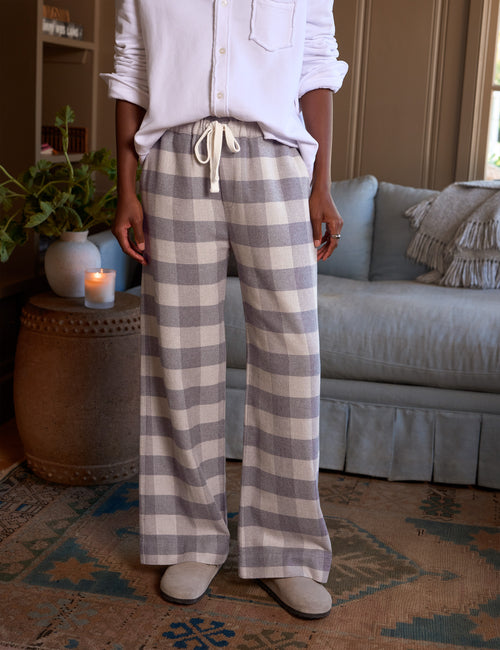 A person standing in a living room wearing wide-leg plaid lounge pants in a light gray and off-white buffalo check pattern, paired with a white button-up shirt and light-colored slippers., front view
