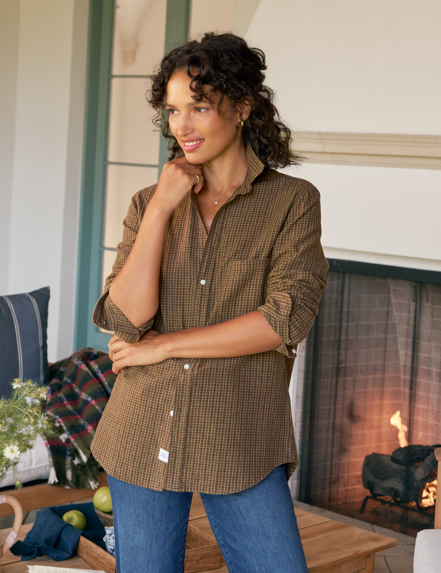 Woman wearing a brown plaid shirt and blue jeans standing in a cozy room with a fireplace.
