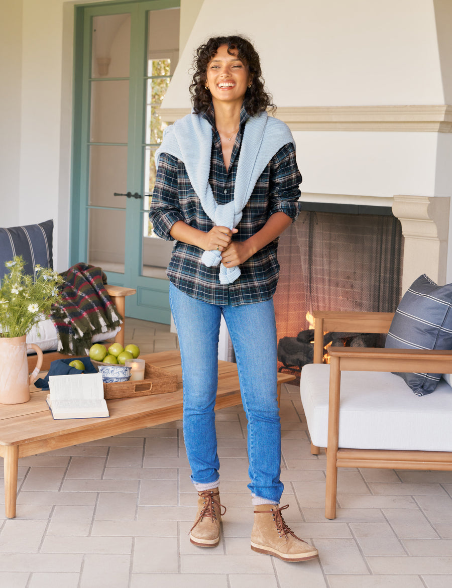 Woman standing in a cozy living room with a fireplace, wearing a plaid shirt, blue jeans, and tan boots.