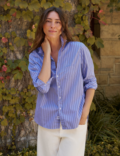 A woman wearing a blue and white vertically striped button-down shirt with white pants, standing outdoors in front of a stone wall and leafy vines., front view