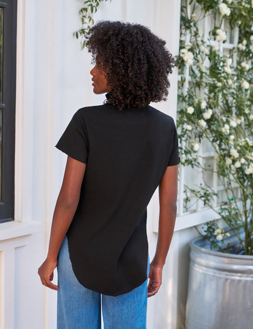 Person wearing a black t-shirt and blue jeans standing in front of a white wall with greenery., alternate view