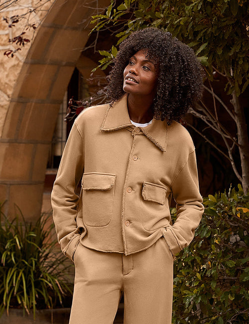 A woman stands outdoors on a stone walkway in front of a stone building with arches and greenery, wearing a tan matching jacket and wide-leg pants set., alternate view
