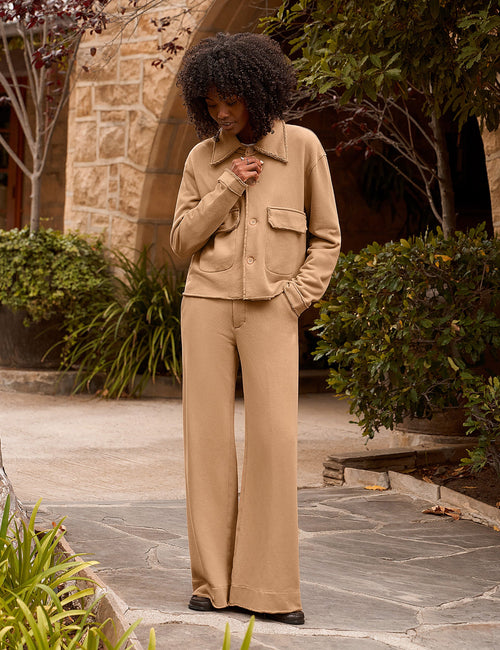 A woman stands outdoors on a stone walkway wearing a matching beige jacket and wide-leg pants set. The setting features stone walls, greenery, and trees in the background., view 4