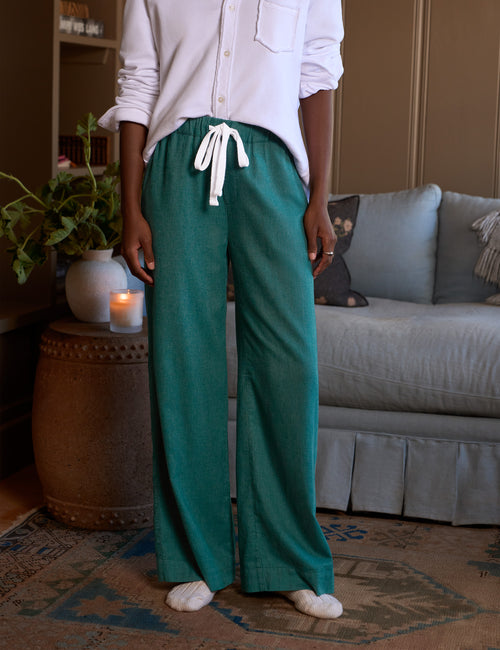 A person standing indoors wearing loose teal-green drawstring pants with a white button-up shirt and white socks, posed in front of a sofa and side table., front view