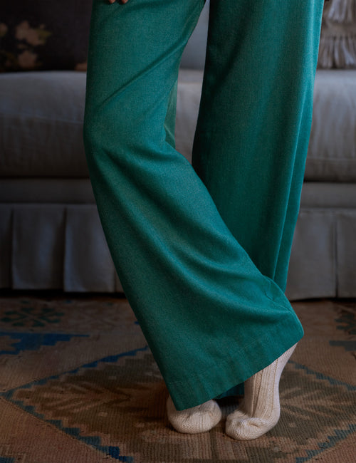 A person wearing teal wide-leg knit pants and cream knit socks, standing on a patterned rug in front of a bed., view 3