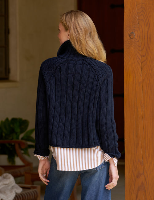 A woman viewed from behind wearing a navy blue ribbed knit mock-neck sweater over a cream and red striped shirt and blue jeans, standing indoors next to a wooden pillar., alternate view