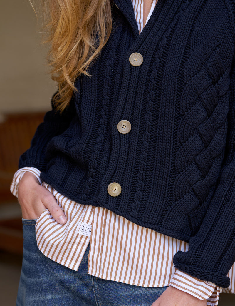 A close-up of a person wearing a navy blue cable-knit cardigan over a cream and orange striped button-down shirt, paired with blue jeans.