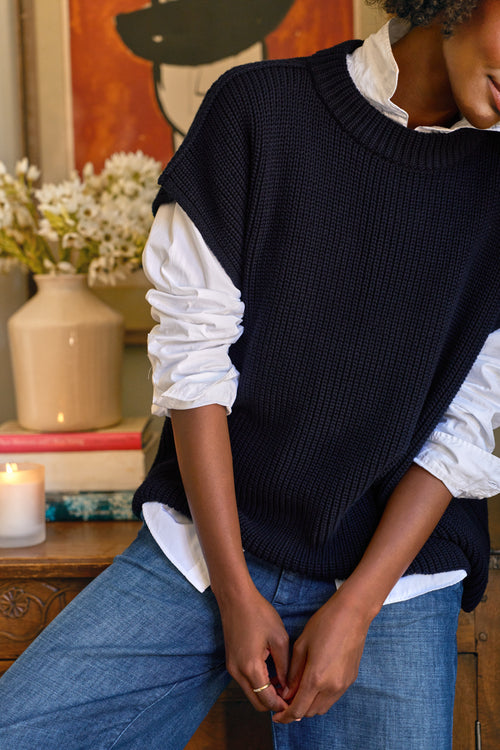 Person wearing a black sweater vest over a white shirt with blue jeans, sitting indoors., alternate view
