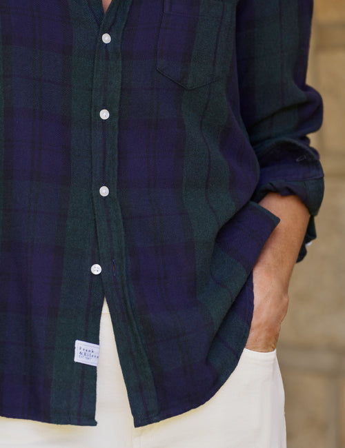 A close-up of a person wearing a dark green and navy plaid button-up shirt with white pants, with one hand in their pocket., view 3