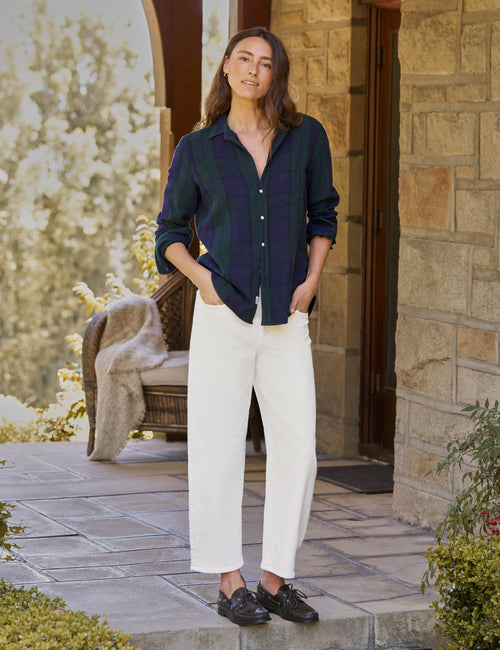 A woman stands on a stone porch wearing a dark navy and green plaid button-up shirt, white cropped pants, and black shoes., view 4