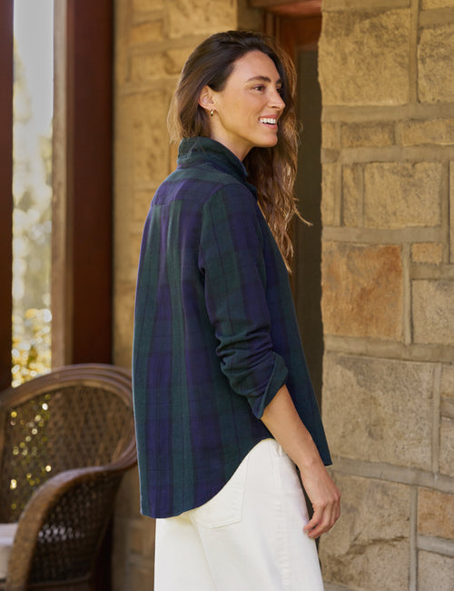 A woman wearing a dark green and navy plaid button-down shirt with white pants, standing on a stone porch next to a wicker chair., alternate view