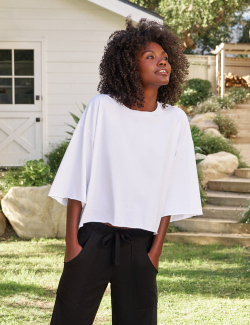 A woman stands outdoors on stone steps in a garden setting, wearing a loose white short-sleeve top and black wide-leg cropped pants with a drawstring waist, paired with black slide sandals., front view