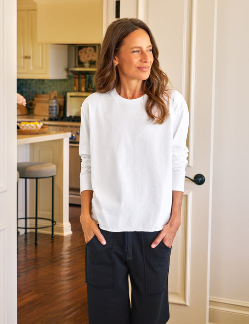 A woman wearing a white crewneck long-sleeve knit tee and dark cargo pants, standing in a doorway of a home interior with a kitchen visible in the background., view 4
