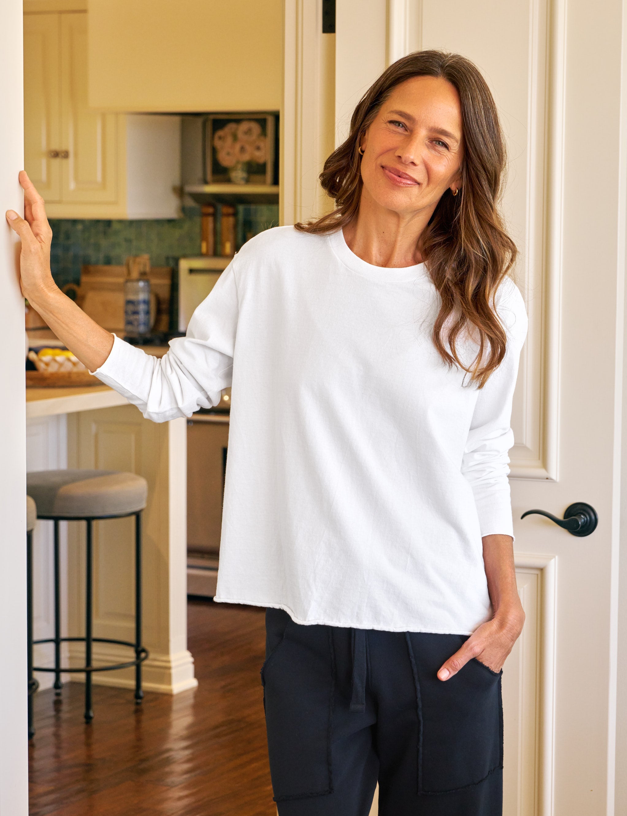 Woman in a white long sleeve tee and navy pants standing in a kitchen.