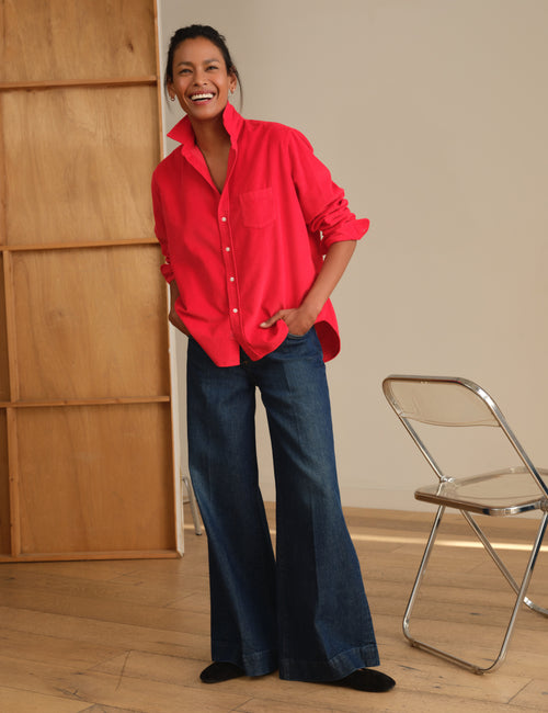 A woman stands in a room wearing an oversized bright red button-up shirt and dark blue wide-leg jeans, with a wooden cabinet on the left and a folding chair on the right., view 6
