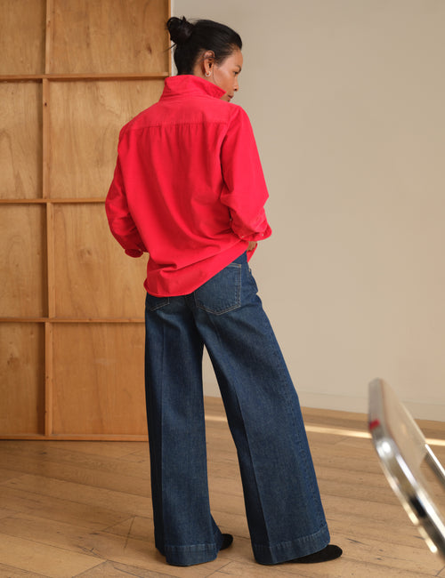 A woman stands with her back to the camera wearing a bright red button-up shirt and dark blue wide-leg denim jeans in a minimalist room with light wood paneling., view 7