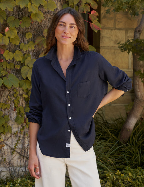 A woman wearing a dark navy blue long-sleeve button-down shirt and white pants, standing outdoors in front of a stone wall and green ivy., front view