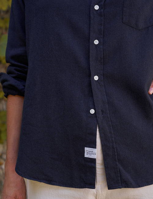Close-up of a person wearing a dark navy button-front shirt with white buttons and a small white brand patch near the hem, paired with light pants., view 3
