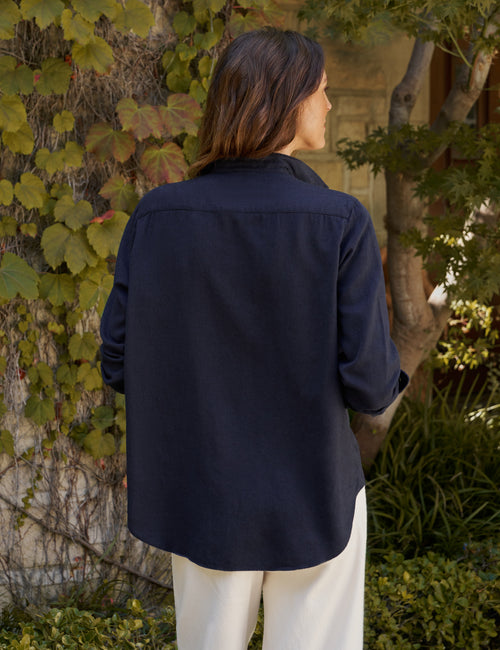 A woman viewed from behind wearing a dark navy blue long-sleeve button-up shirt and off-white trousers, standing outdoors in front of a wall covered in green ivy., alternate view