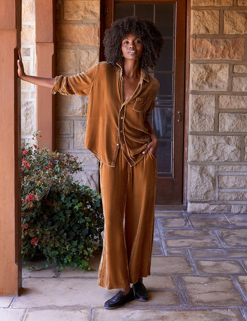 A woman stands on a stone porch wearing a matching two-piece set in a gold velvet fabric. The set includes a button-front top and wide-leg pants, styled with black loafers., front view