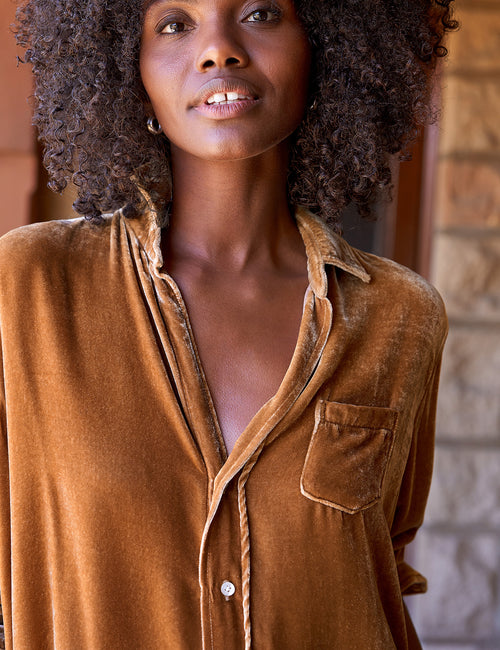 A close-up shot of a woman with curly dark hair wearing a gold velvet button-down shirt with a chest pocket, set against a warm stone wall background., alternate view