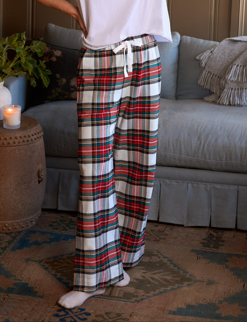 A woman stands in a living room wearing a white short-sleeve t-shirt and high-waisted plaid lounge pants with a white drawstring. The pants feature a red, green, and black plaid pattern on a white base., front view