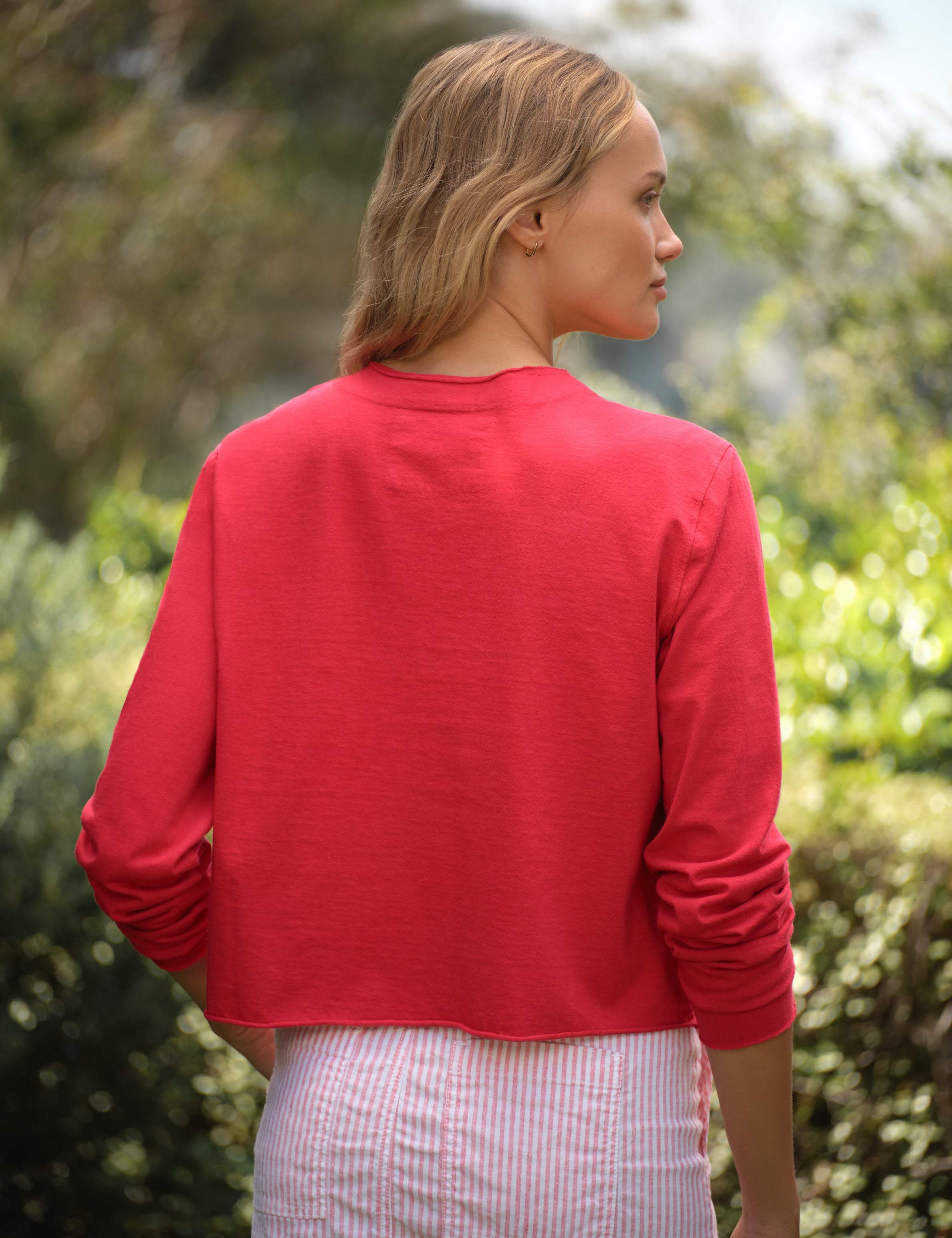 Woman wearing a red cardigan and striped skirt standing outdoors with greenery in the background, view 4