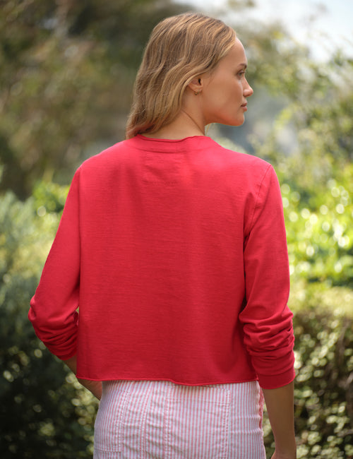 Woman wearing a red cardigan and striped skirt standing outdoors with greenery in the background, view 4