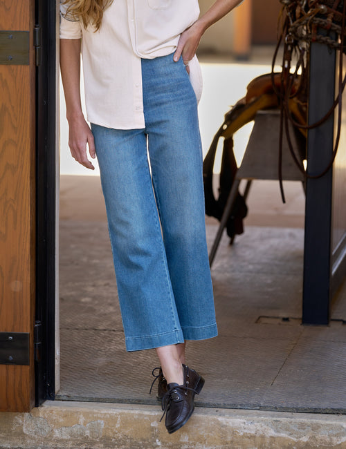 A person wearing wide-leg cropped blue denim jeans, a cream blouse, and dark brown leather shoes, standing in a doorway., front view