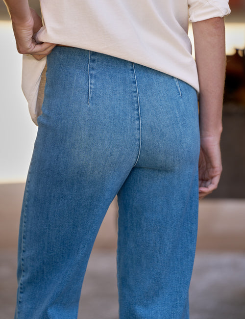 Close-up back view of a person wearing light-to-medium blue denim jeans and a cream-colored top, standing in an indoor setting., view 3
