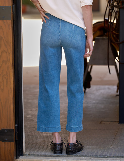A person shown from the back wearing light-to-medium blue wide-leg cropped jeans, a cream top, and black shoes, standing in a doorway., alternate view