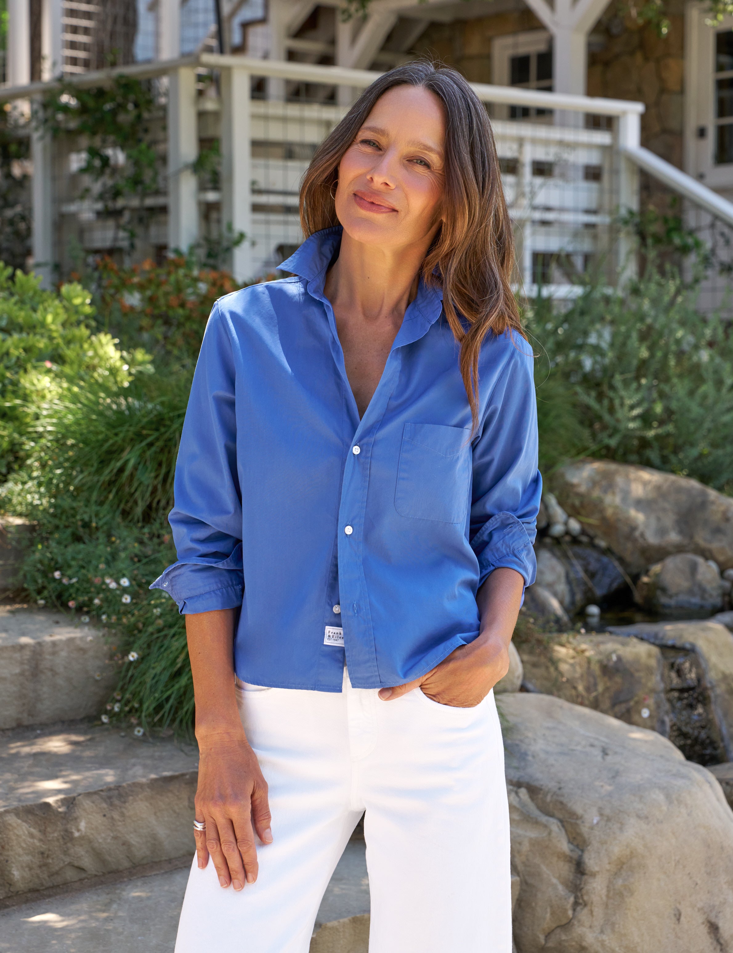 Woman wearing a blue shirt and white pants standing outdoors with greenery and a building in the background, front view