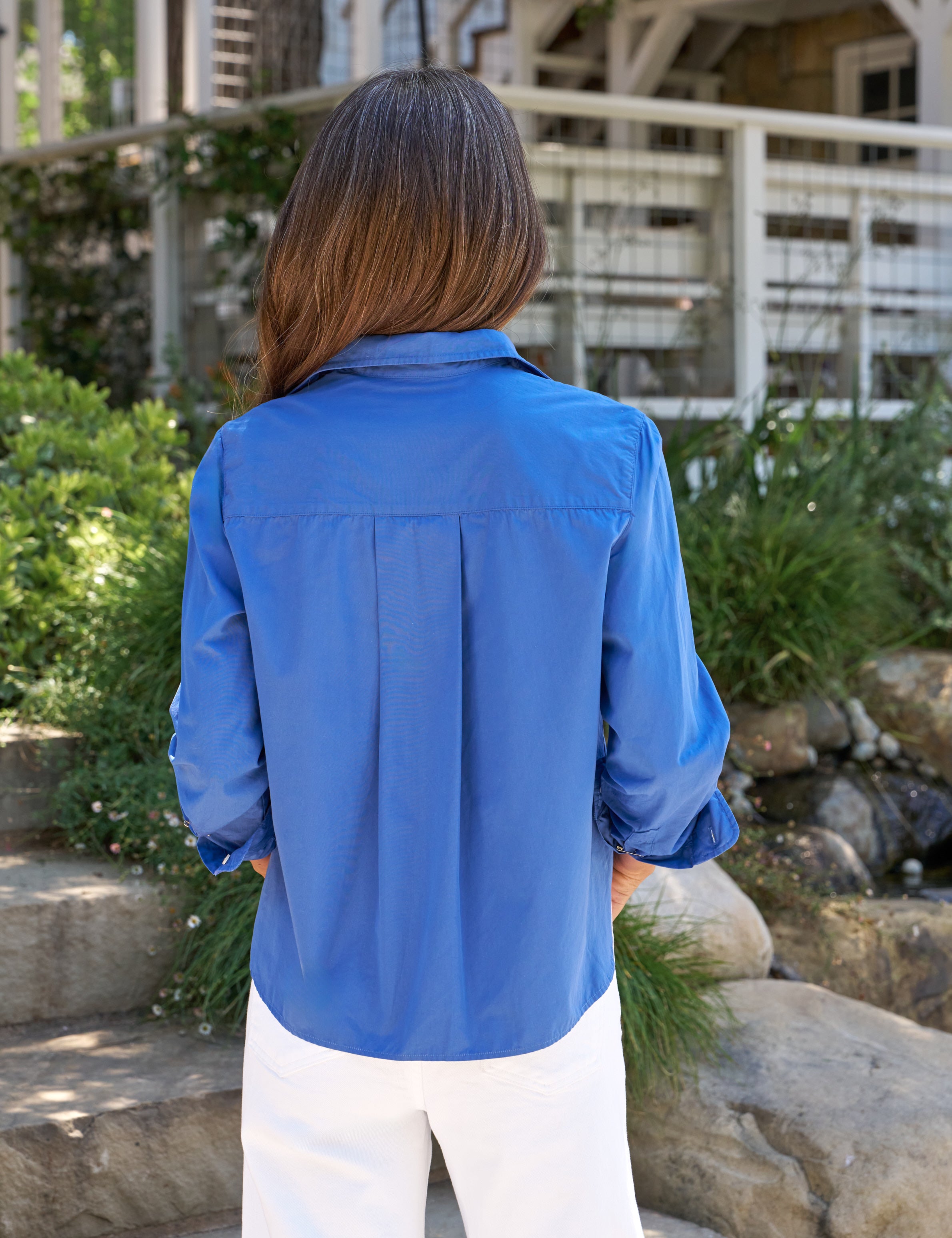 Person wearing a blue shirt standing outdoors with greenery and a building in the background, alternate view