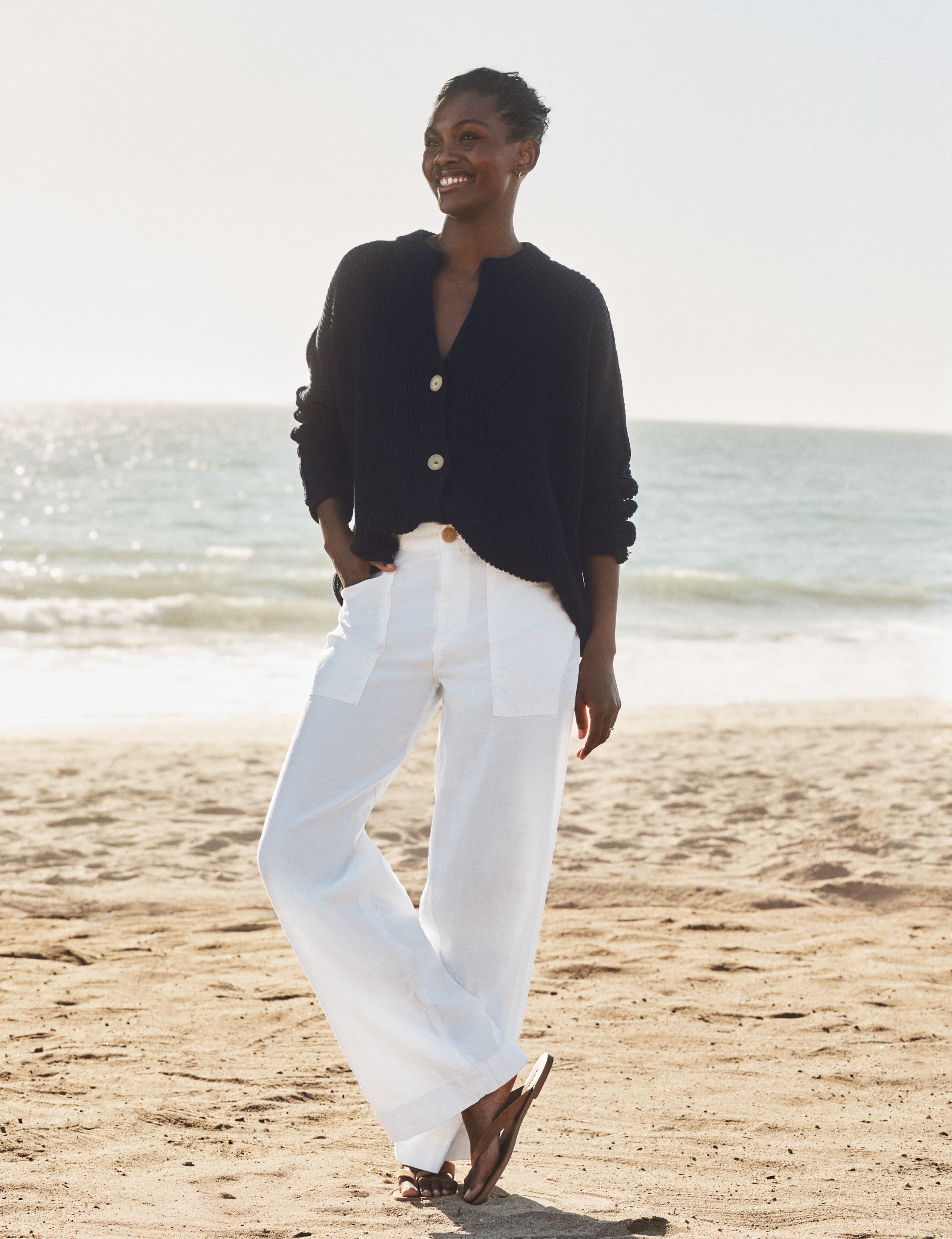 Woman standing on a beach wearing a navy cardigan and white pants., view 4