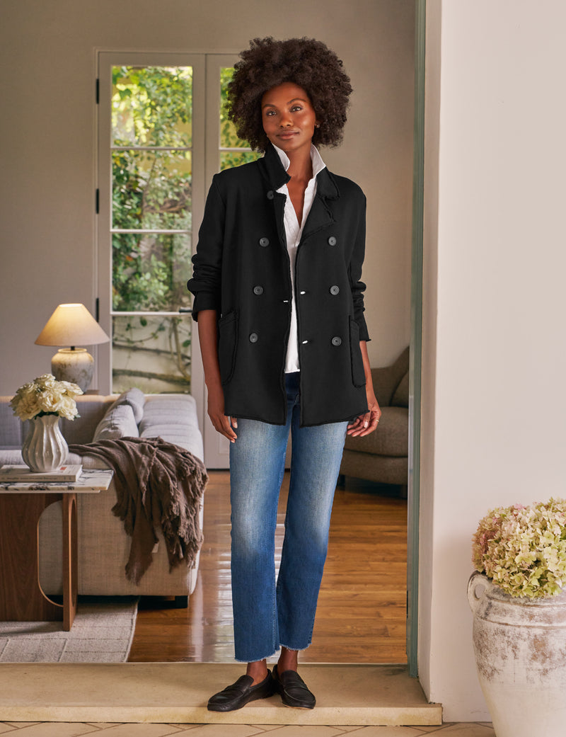 Woman standing in a home interior wearing a black coat and blue jeans.