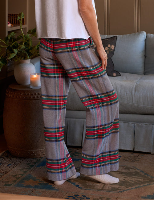 A woman stands in a living room wearing a white short-sleeve top and gray plaid lounge pants with red, green, and black lines. The room has brown paneled walls, a light blue sofa, and a patterned rug., alternate view