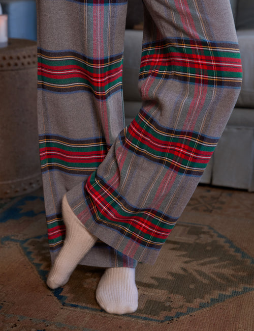 Close-up of a person wearing gray wide-leg plaid pajama pants with red, green, and blue stripes, paired with white socks, standing on a patterned rug., view 3