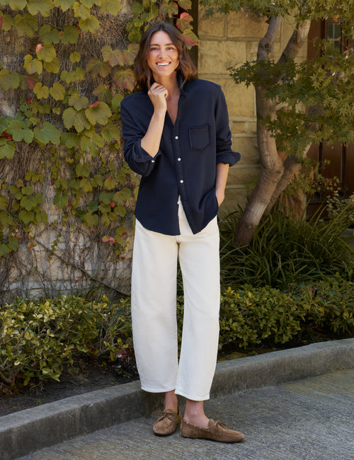 A woman stands outdoors in front of a stone wall covered in green vines, wearing a dark navy button-up shirt, off-white wide-leg pants, and brown suede loafers., view 4