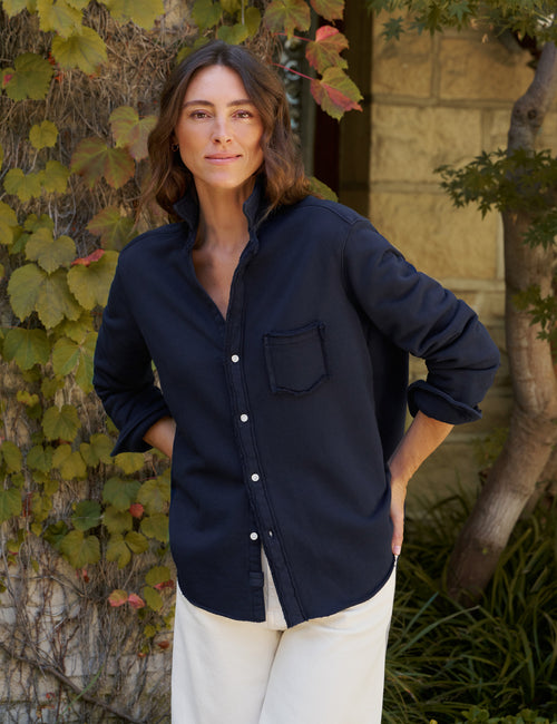 A woman wearing a dark navy blue long-sleeve button-up shirt with a chest pocket, paired with off-white pants, standing outdoors in front of a stone wall and leafy vines., front view