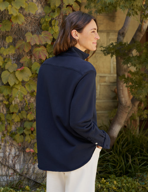 A woman wearing a dark navy blue long-sleeve shirt and white pants, standing outdoors in front of a stone wall covered in green and autumn-colored ivy., alternate view