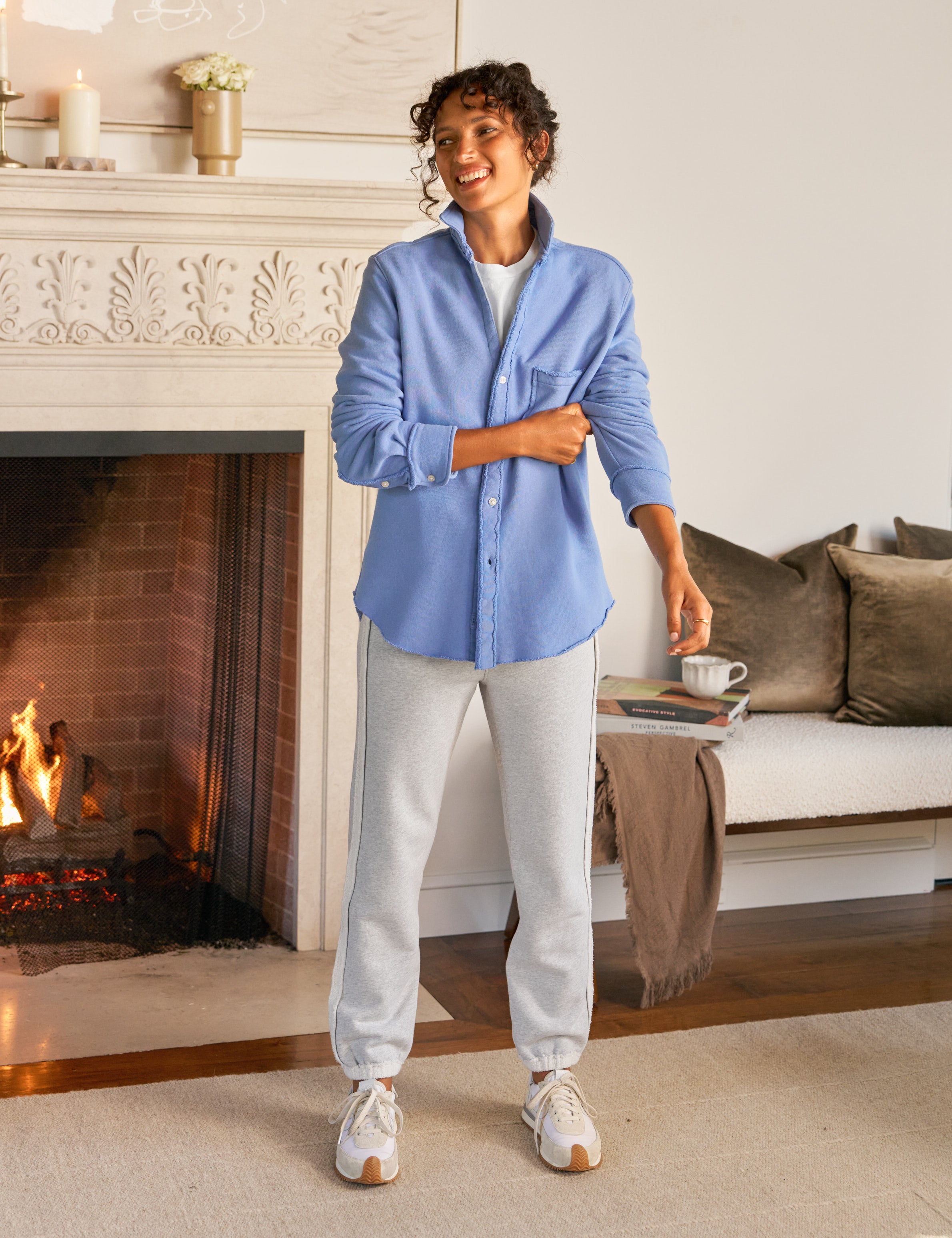 Woman standing in a cozy living room with a fireplace, wearing a blue shirt and light gray pants.