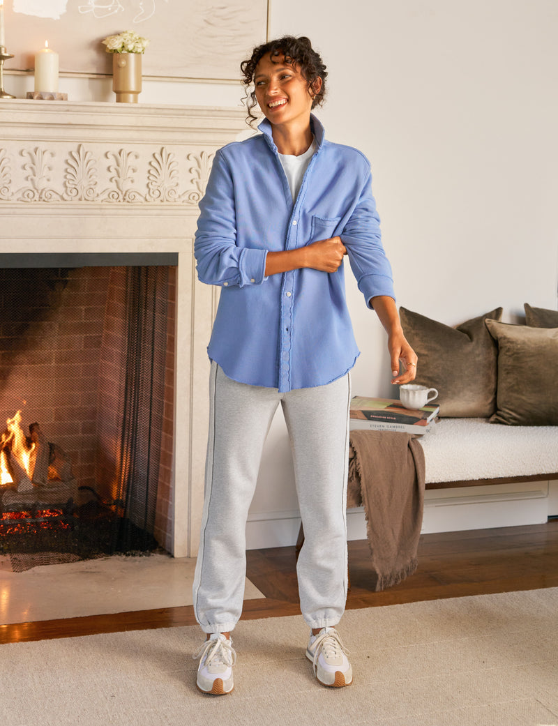 Woman standing in a cozy living room with a fireplace, wearing a blue shirt and light gray pants.