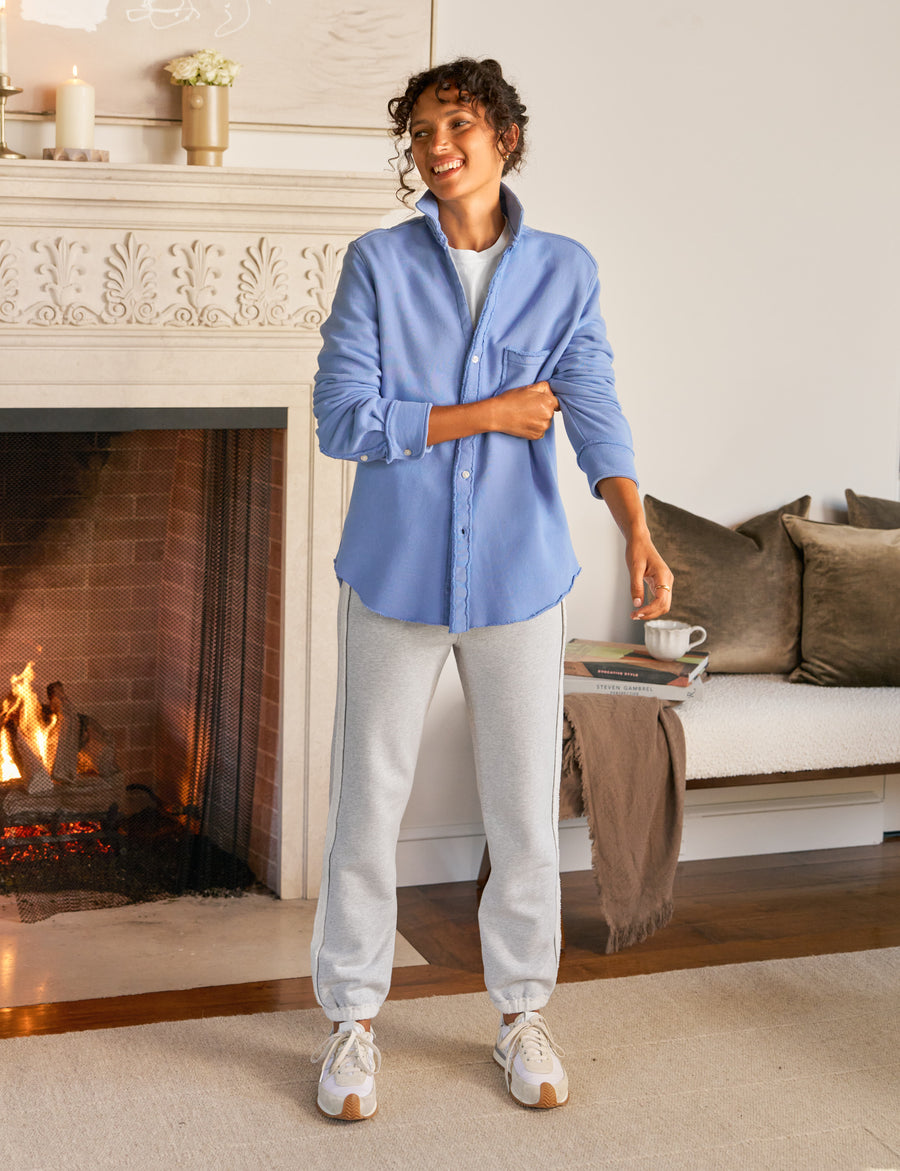 Woman standing in a cozy living room with a fireplace, wearing a blue shirt and light gray pants.