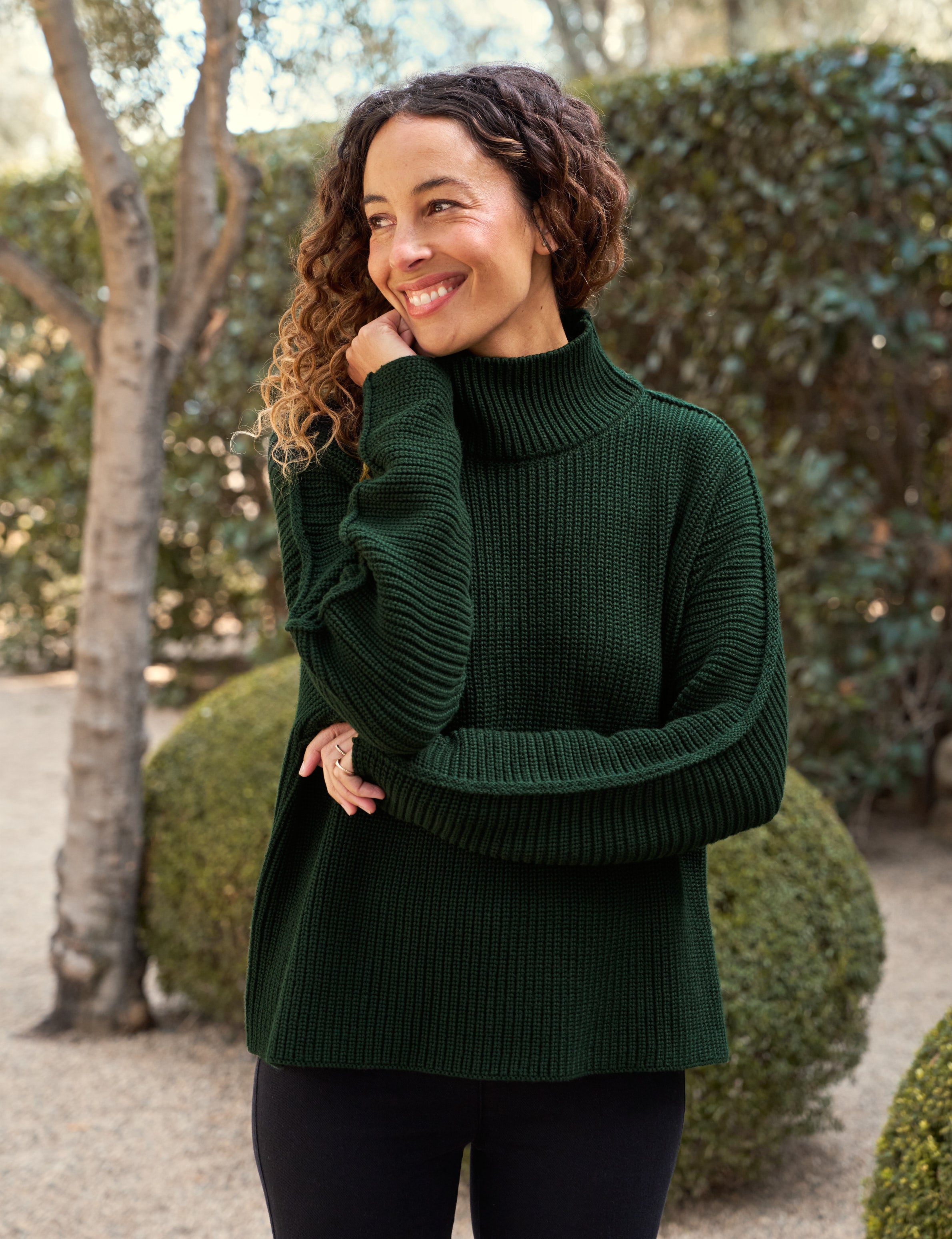Woman wearing a green sweater standing outdoors with trees and shrubs in the background
