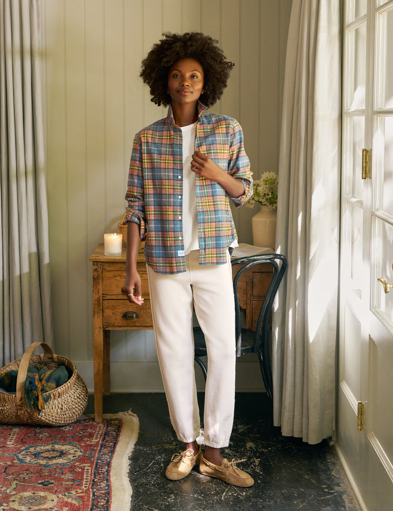 Woman wearing a plaid shirt and white pants standing in a room with a desk and window.