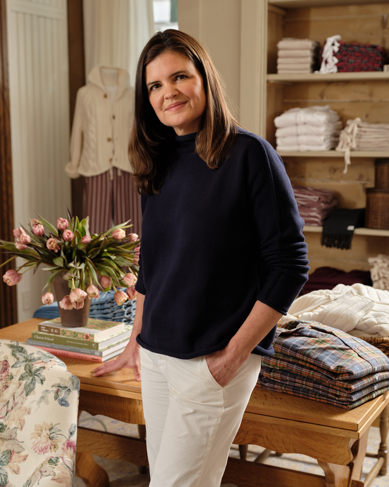 Woman standing in a room with shelves displaying folded clothes and a table with flowers and books.