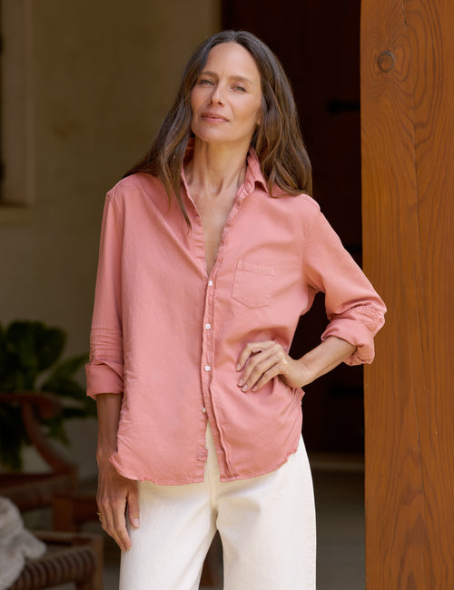 A woman wearing a dusty pink long-sleeve button-up shirt and white pants, standing in a warm-toned indoor setting with a wooden door frame., front view
