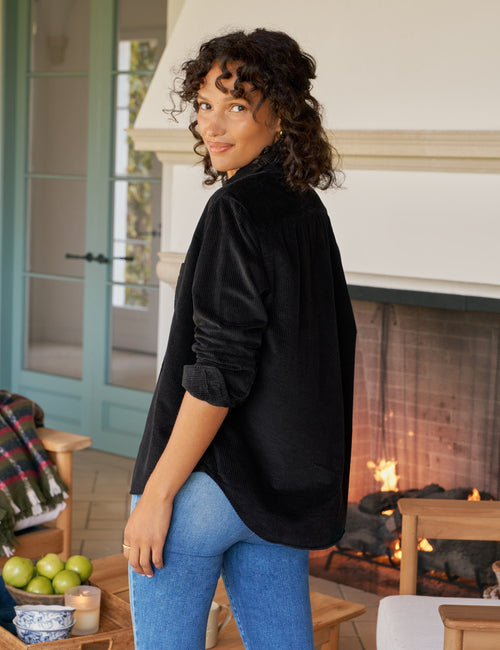 Woman wearing a black shirt and blue jeans standing in a cozy living room with a fireplace., alternate view