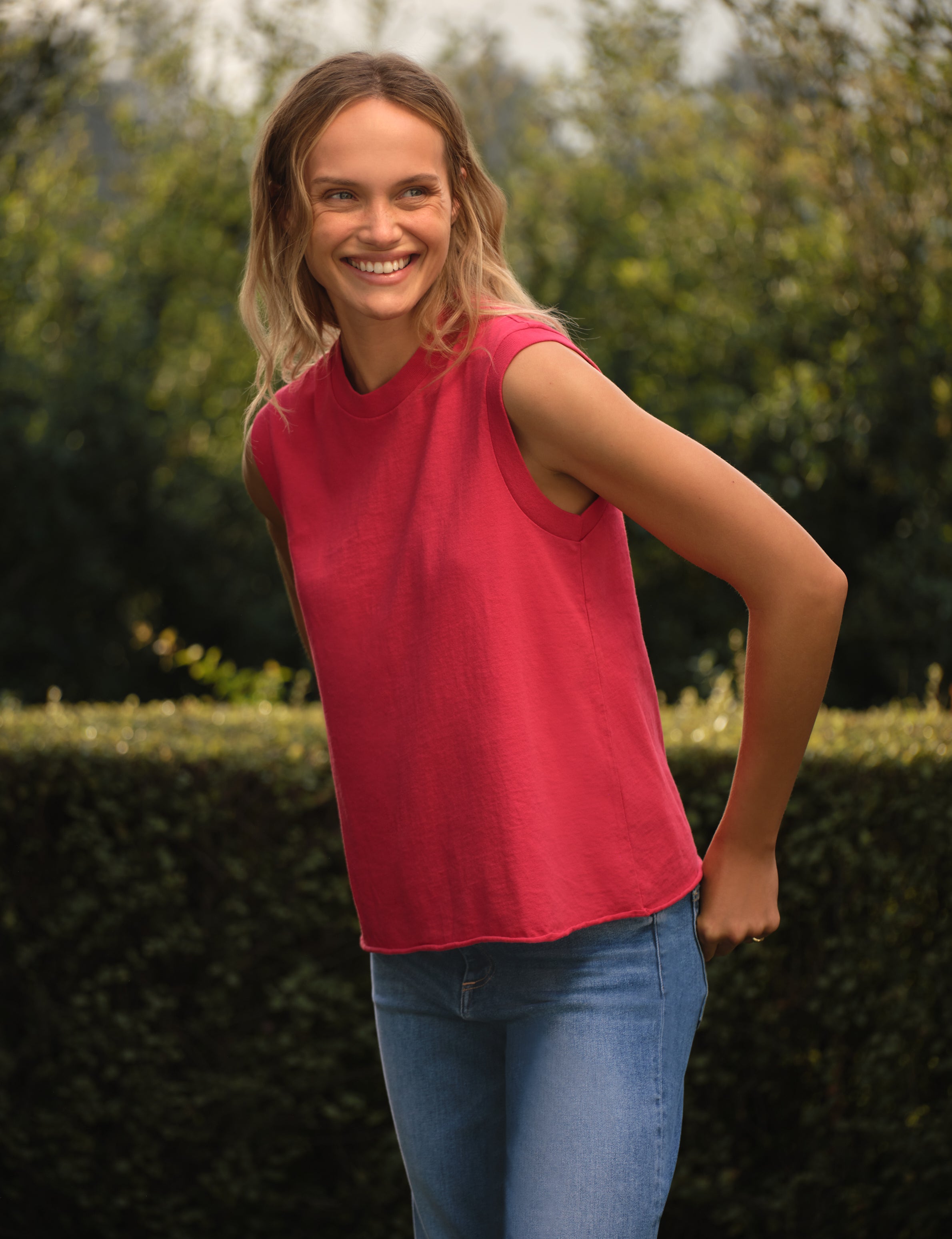 Woman wearing a red sleeveless top and blue jeans standing outdoors with greenery in the background, front view